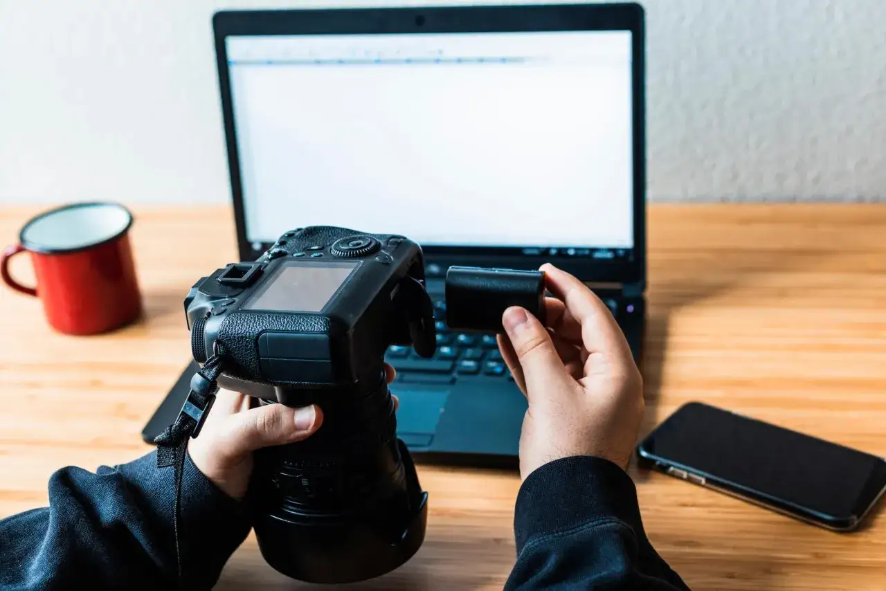 Ręce wkładają baterię do aparatu Canon EOS. Obok leży laptop, smartfon i czerwony kubek.