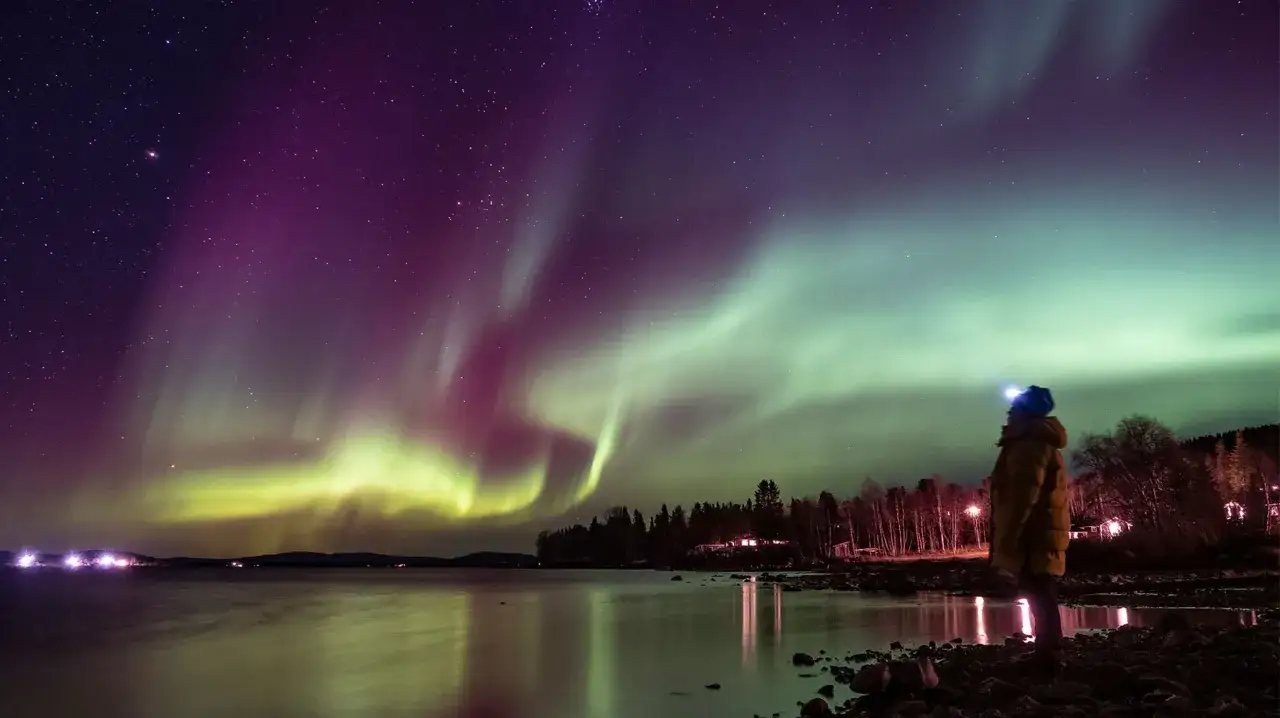 En person ser norrsken &ouml;ver en sj&ouml; n&auml;ra Kiruna i Lappland.