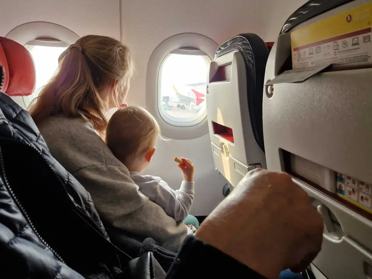 Dziecko na kolanach mamy patrzy przez okno samolotu. Wizzair od ilu lat można latać samemu? To pytanie nurtuje wielu młodych podróżników.