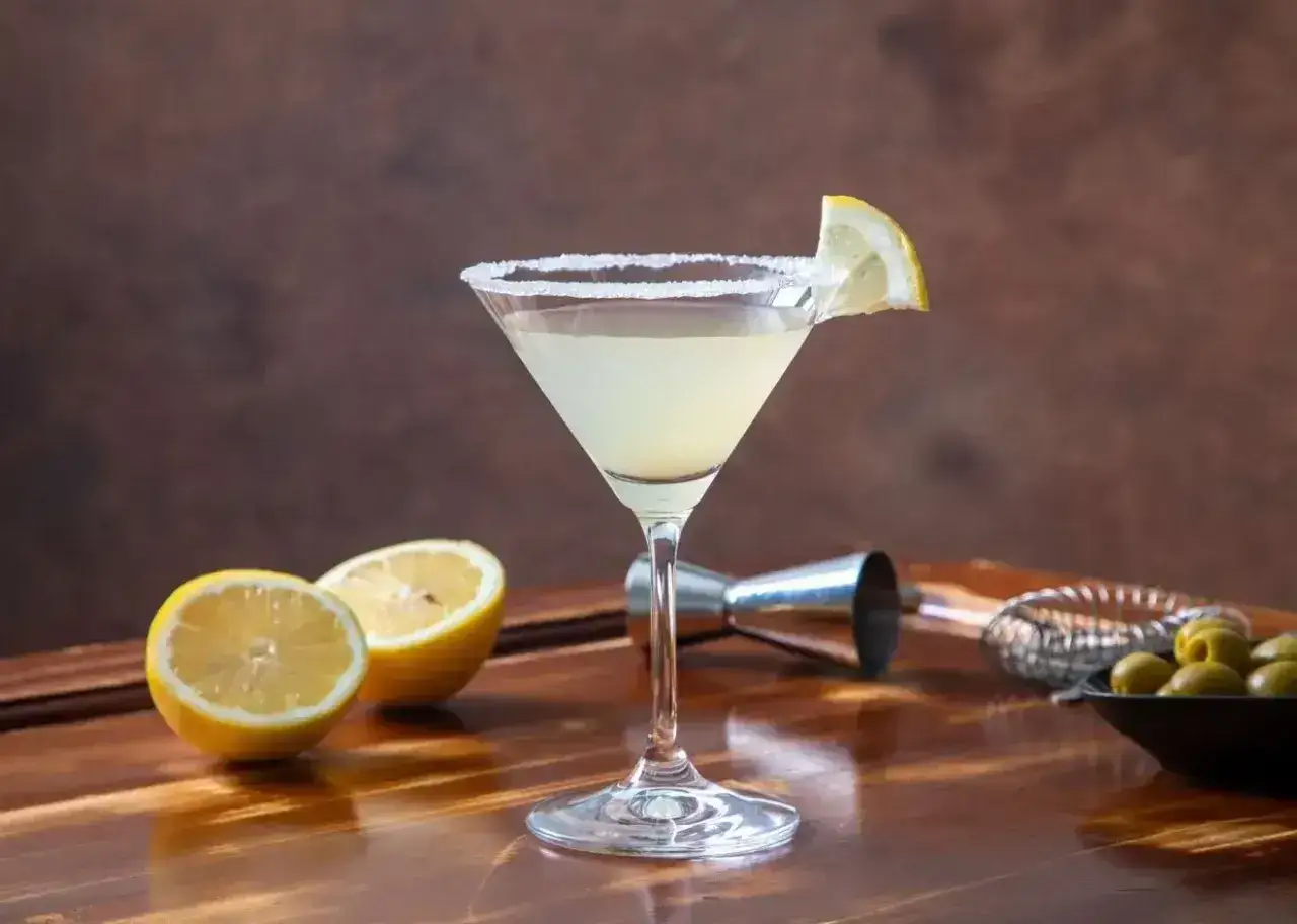 Orzeźwiający martini ze spritem, udekorowany plasterkiem cytryny i solnym brzegiem kieliszka. Obok leżą połówki cytryn i oliwki.
