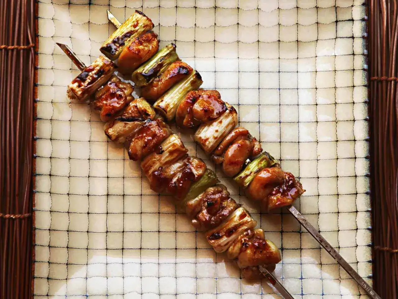 Dwa szaszłyki yakitori z kurczakiem i zieloną cebulką, polane słodkim sosem, leżą na talerzu z siatką.