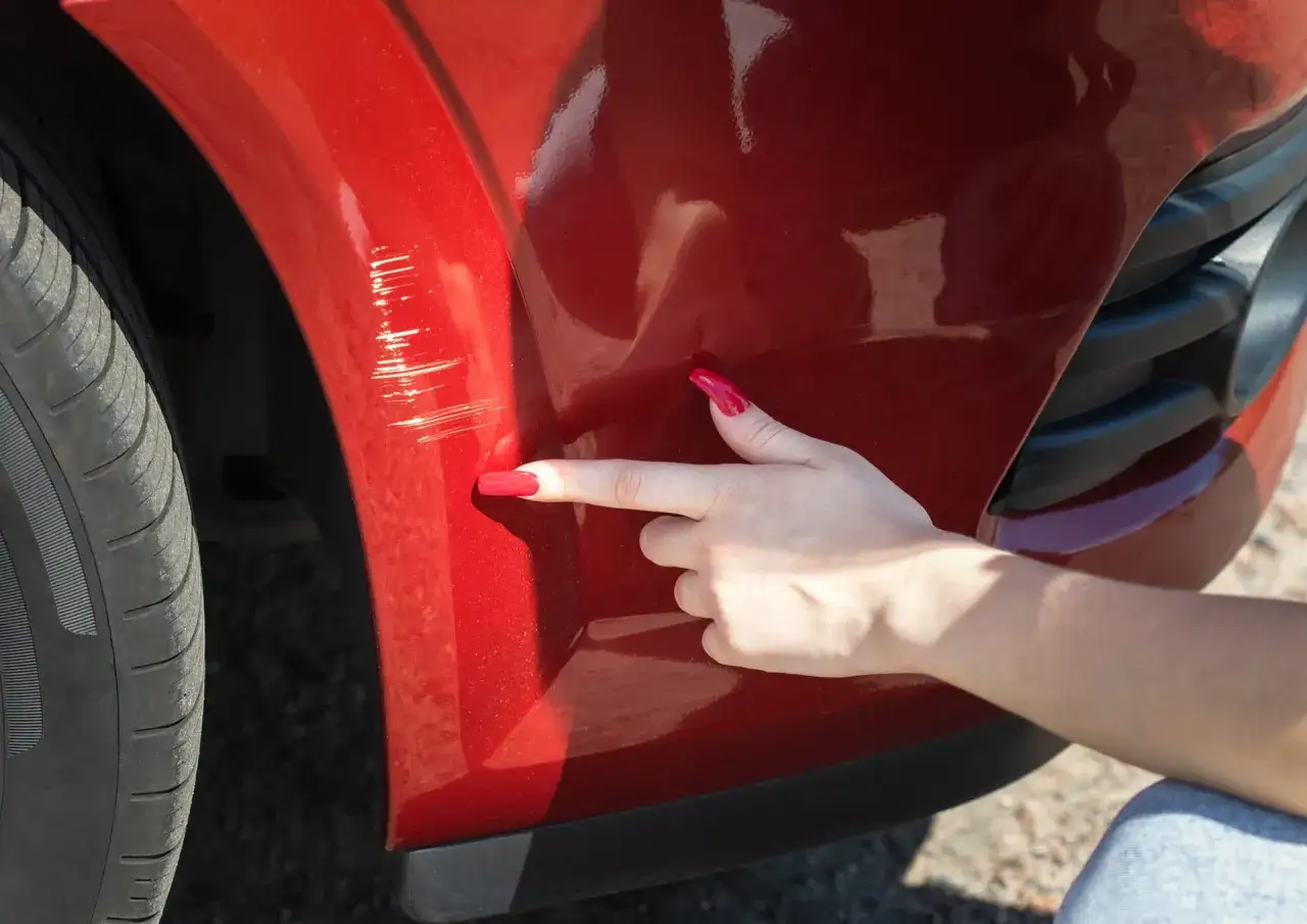 A hand with bright red nails points to deep scratches on a red car bumper. Wondering if you can buff out scratches like these?