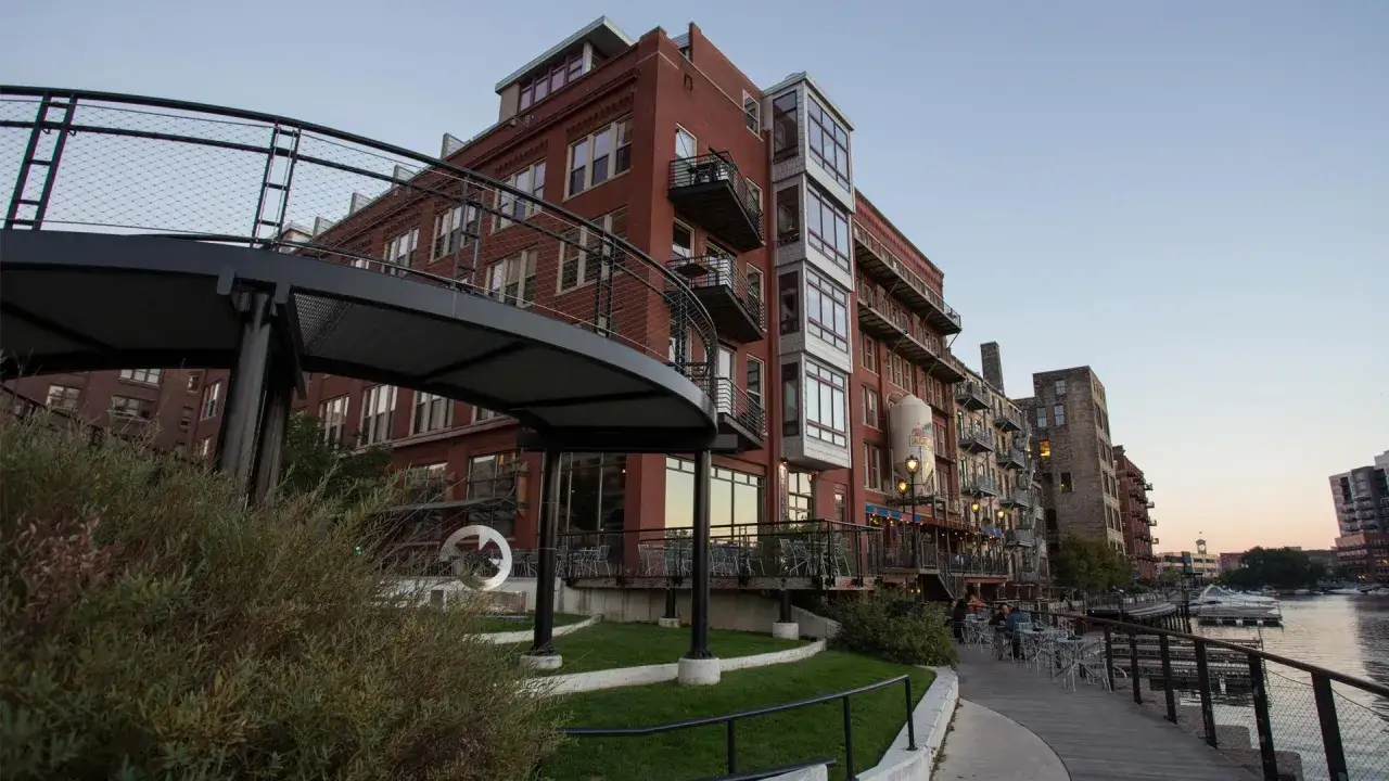 A perfect weekend in Milwaukee: brick buildings line a riverwalk with outdoor seating, boats docked nearby, and a modern spiral walkway.