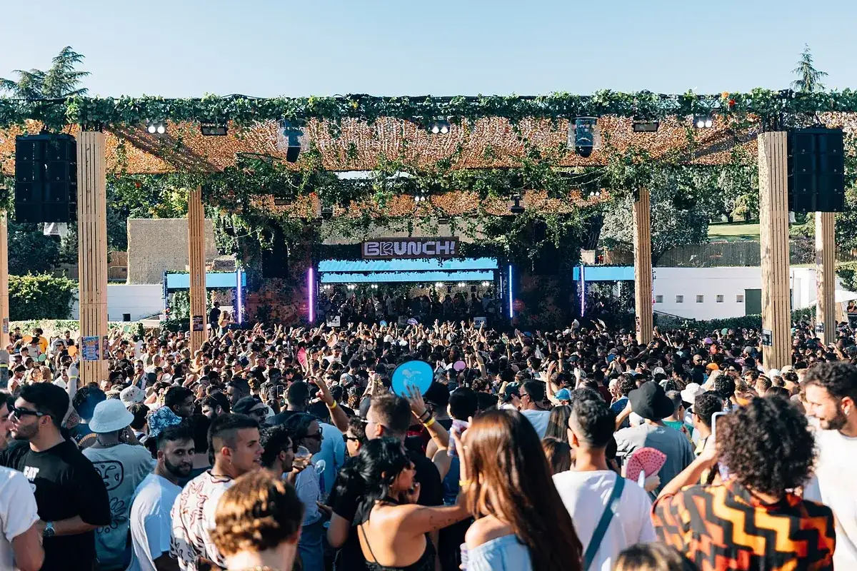 Multitud disfrutando de un d&iacute;a soleado en Brunch Electronik Lisboa, con un escenario decorado con vegetaci&oacute;n y luces vibrantes.
