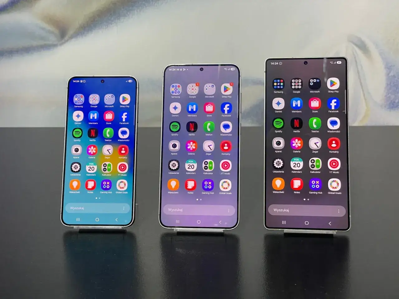Trzy smartfony z różnymi **markami telefonów** prezentują swoje ekrany z ikonami aplikacji, w tym Samsung, Google i Facebook.