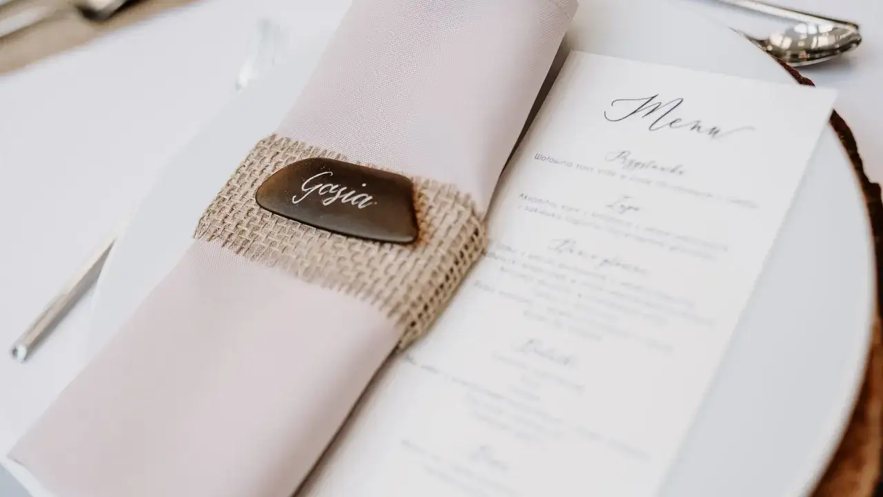 Eleganckie ręczne wypisywanie zaproszeń ślubnych. Na talerzu leży r&oacute;żowa serwetka z lnianym pierścieniem i tabliczką 