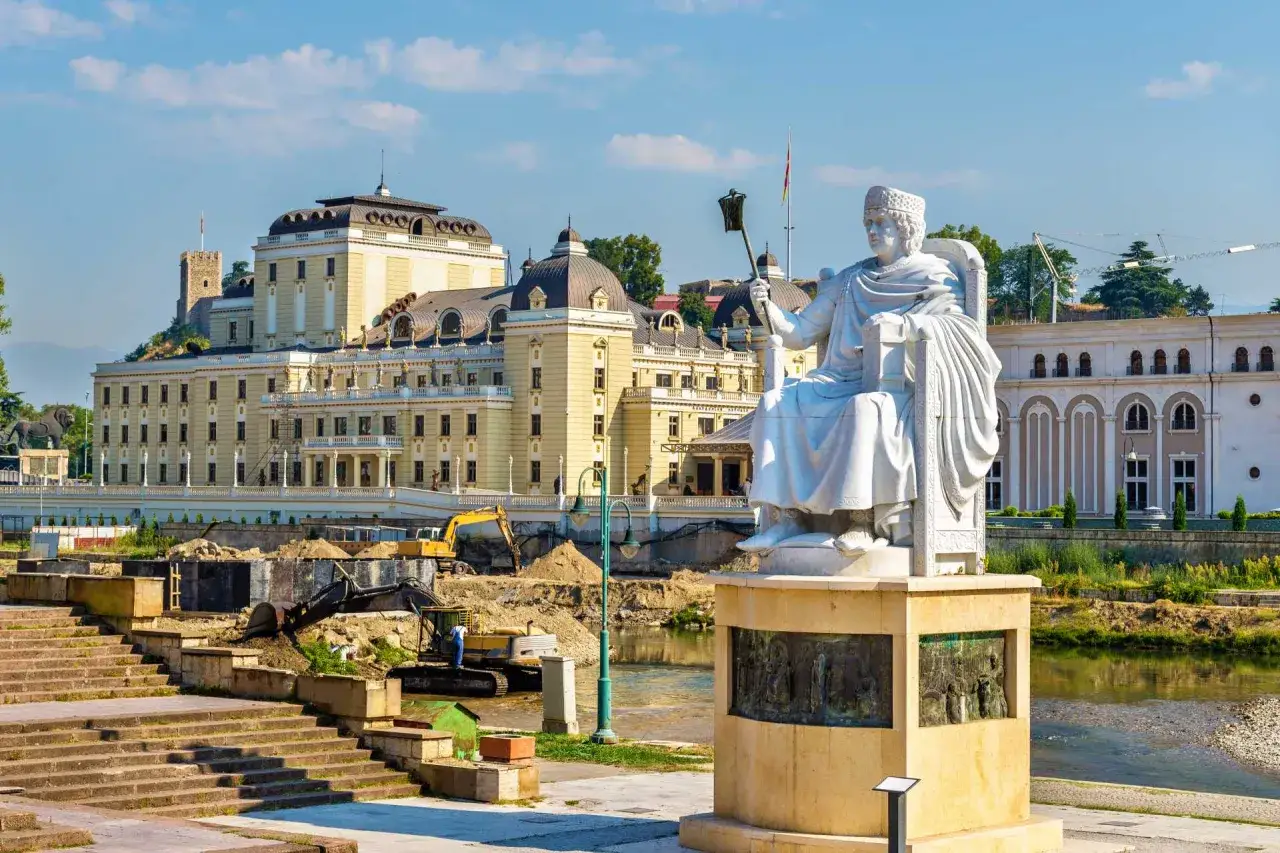 Skopje co zobaczyć: monumentalny pomnik siedzącego władcy na tle okazałych budynków i placu budowy nad rzeką.