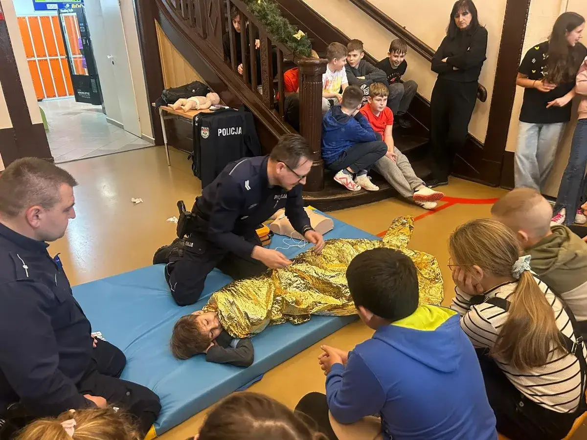 Policjanci prowadzą pokaz pierwszej pomocy, ucząc dzieci edukacji dla bezpieczeństwa. Na macie leży chłopiec w folii termicznej, a funkcjonariusze demonstrują resuscytację.
