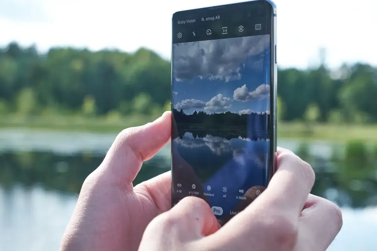 Ręce trzymają smartfon Samsung S10, aparat uchwycił odbicie nieba i drzew w spokojnej tafli jeziora.