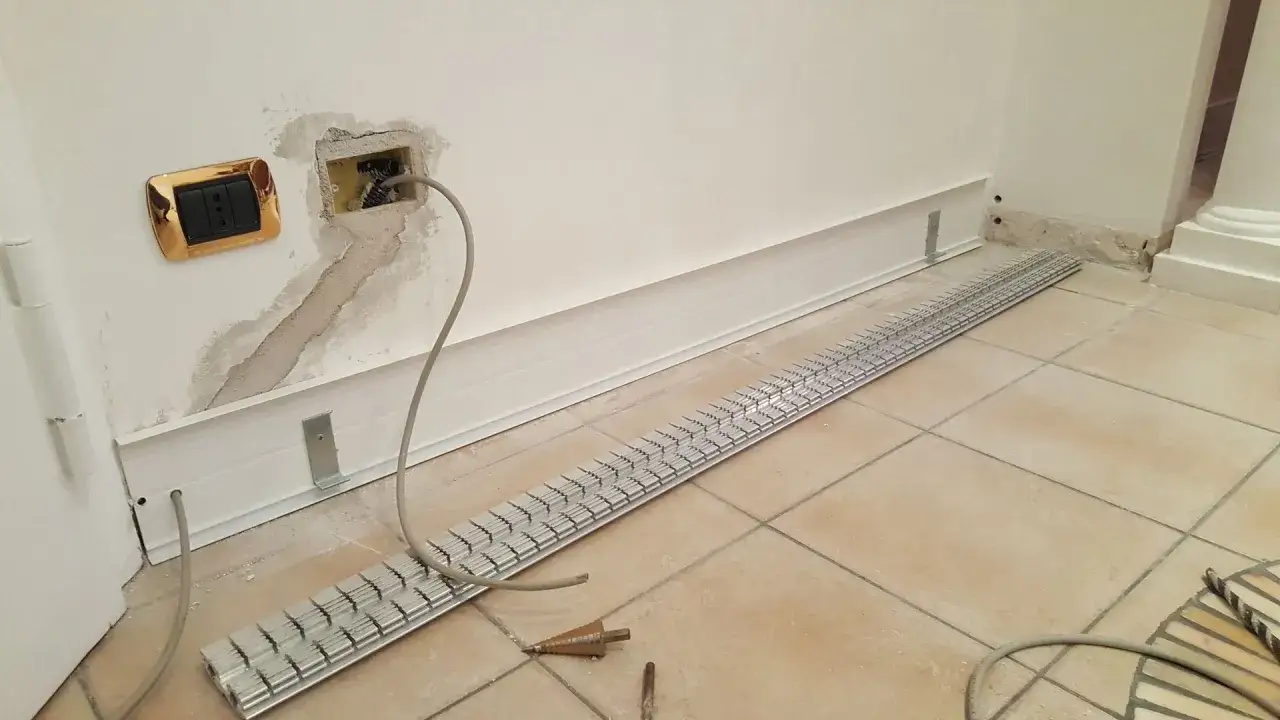 Lavori in corso per collegare fili elettrici. Una presa dorata è visibile su un muro intonacato, con cavi che escono da una scatola. Una griglia metallica è posata sul pavimento piastrellato.