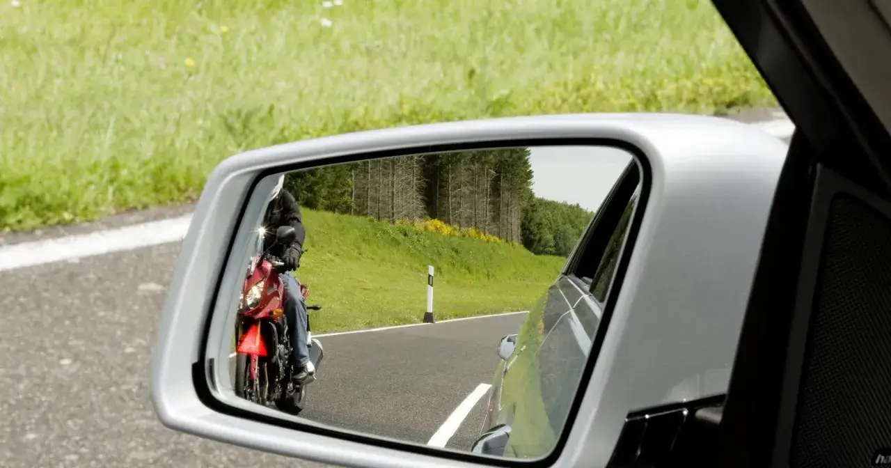 Motocyklista w białym kasku i czarnej kurtce jedzie na czerwonym motocyklu, widoczny w kamerze w lusterku bocznym samochodu.