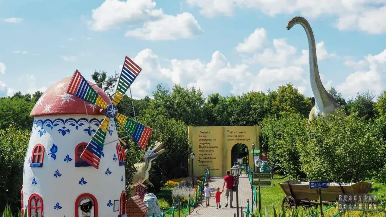 Kolorowy wiatrak i wielki dinozaur w parku dinozaurów Łódź. Rodzina spaceruje ścieżką wśród zieleni.