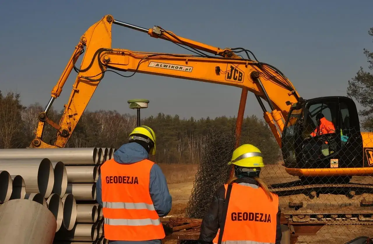 Na budowie geodeci w kamizelkach obserwują pracę koparki. Zastanawiają się, ile kosztuje wynajem koparki do takich prac.