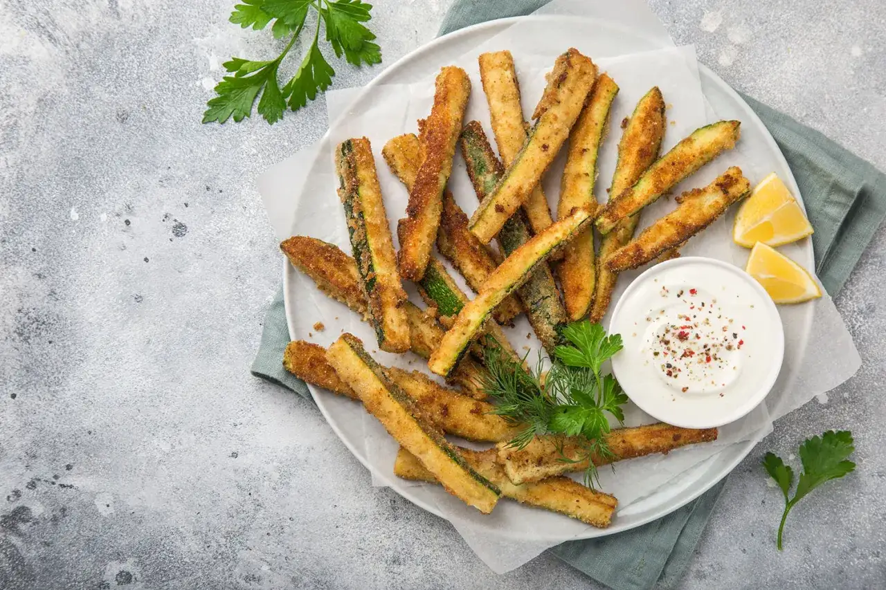 Knusprige frittierte Zucchini-Sticks auf einem Teller mit Dip, Zitrone und Petersilie.
