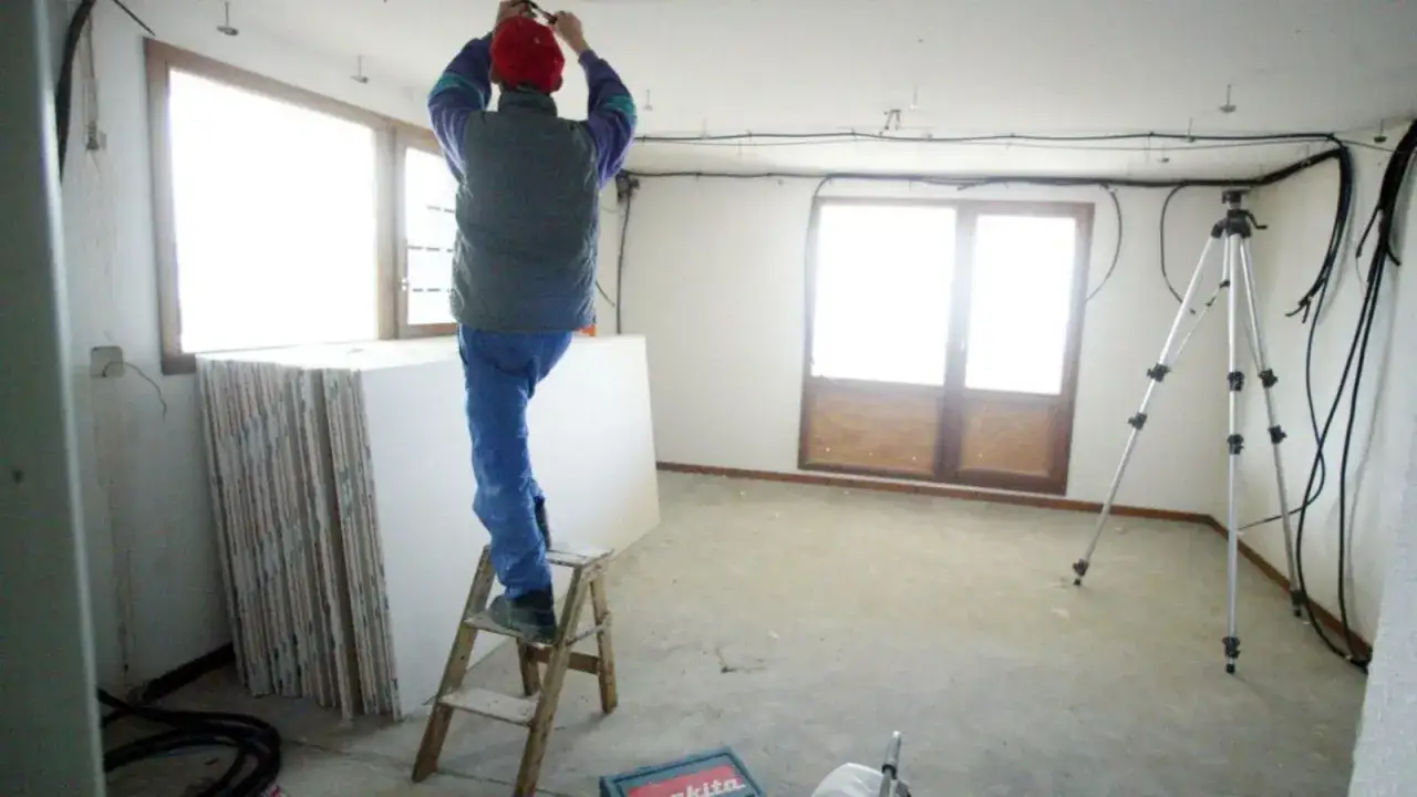 Un ouvrier sur un escabeau installe des câbles au plafond. La pièce est en chantier, avec des plaques de plâtre empilées. Une cuisine mal posée quel recours ?