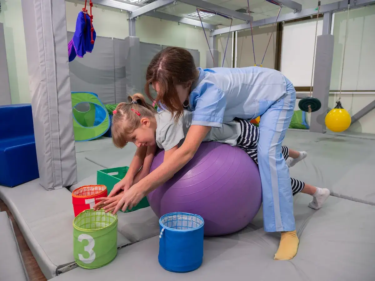 Ni&ntilde;a con par&aacute;lisis cerebral se ejercita en una pelota de fisioterapia, guiada por un terapeuta.