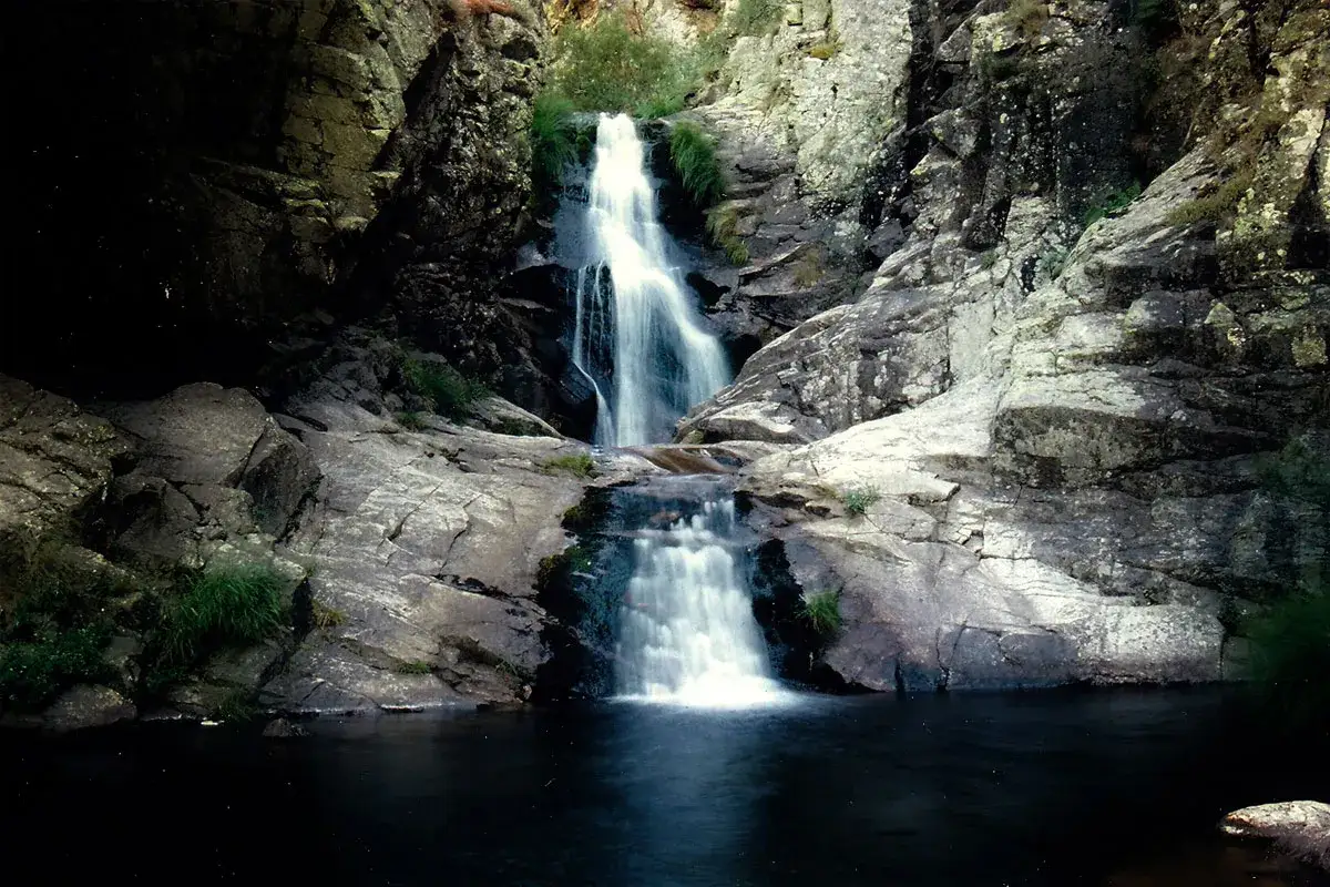 Cascada en el **parque nacional de la Sierra de Guadarrama**. El agua cae sobre rocas y forma una poza oscura.