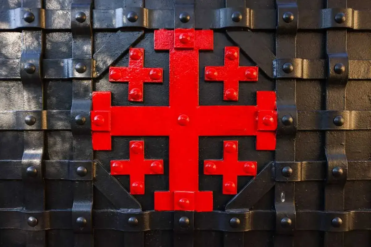 Czerwony krzyż jerozolimski na czarnej, metalowej bramie. Czy katolik może nosić krzyż jerozolimski? Symbol ten jest często noszony przez wiernych.
