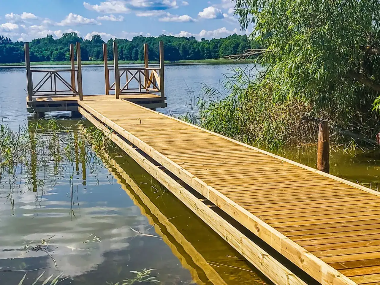 Drewniany pomost prowadzi do prywatnych stawów wędkarskich. Woda odbija błękitne niebo i zielone drzewa.