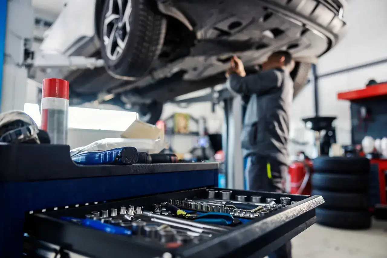 Mechanik pracuje pod samochodem na podnośniku. W tle widać narzędzia i opony. Szybka wymiana oleju to klucz do sprawnego działania auta.