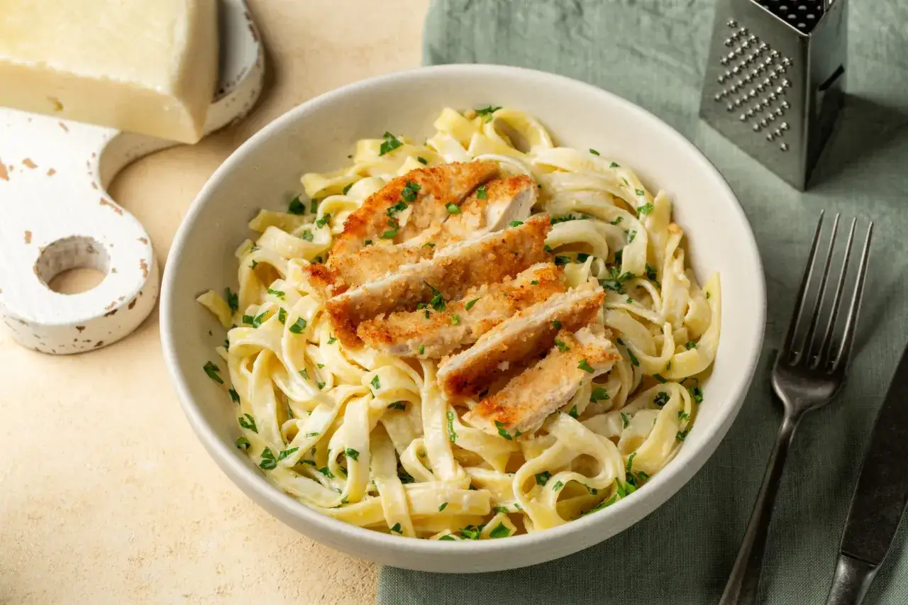 Un plat de fettuccine alfredo cr&eacute;meux, garni de fines tranches de poulet pan&eacute; et de persil frais. Un morceau de fromage et une r&acirc;pe sont visibles &agrave; c&ocirc;t&eacute;.