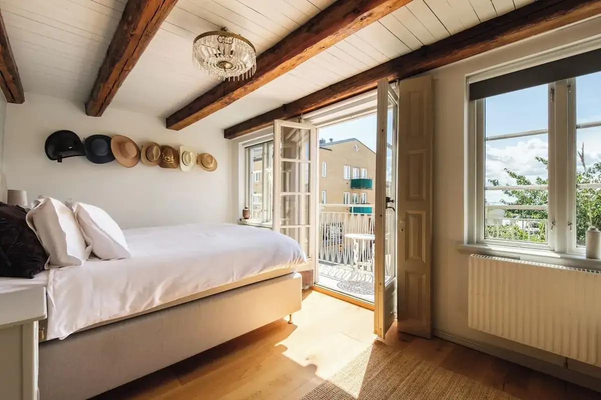 Ett mysigt sovrum med synliga bjälkar och en balkongdörr som leder ut till en solig utsikt. Kanske ett rum på hotellet Höganäs?