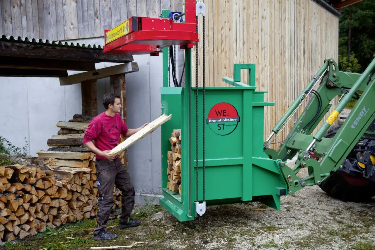 Mann belädt ein grünes **Bündelgerät** mit Holzscheiten. Ein Traktor steht bereit.