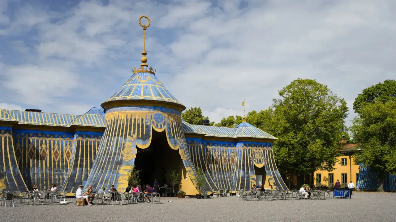 En blågul cirkustältliknande byggnad med guldfärgade detaljer i Haga parken. Människor sitter vid bord utomhus.
