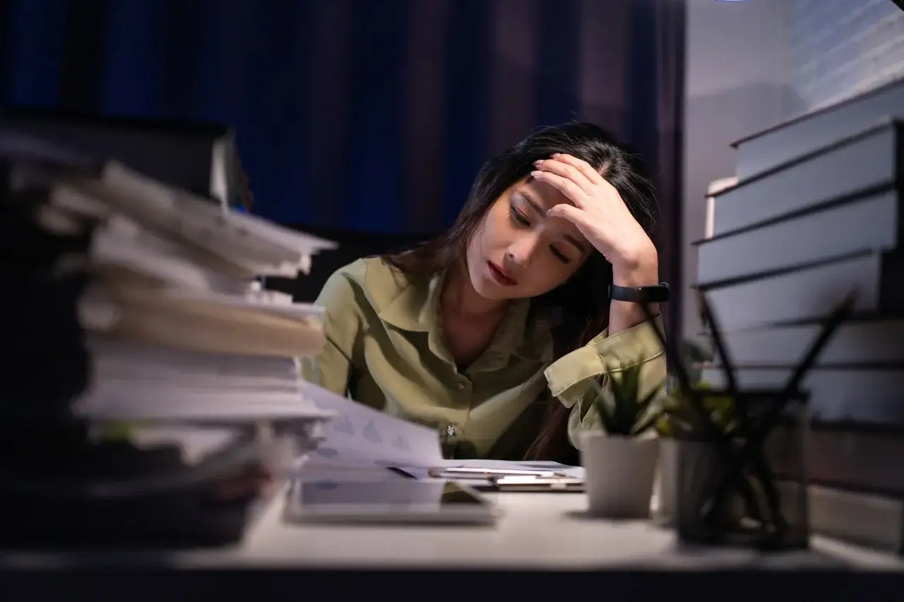 En kvinna med handen mot pannan ser omotiverad på jobbet, omgiven av högar med papper och böcker i svagt ljus.