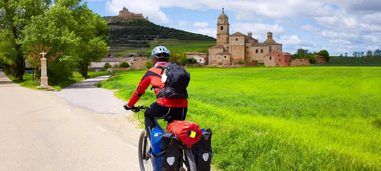 Ciclista en ruta por la EuroVelo 3, con un castillo y una iglesia en el horizonte, rodeado de campos verdes.