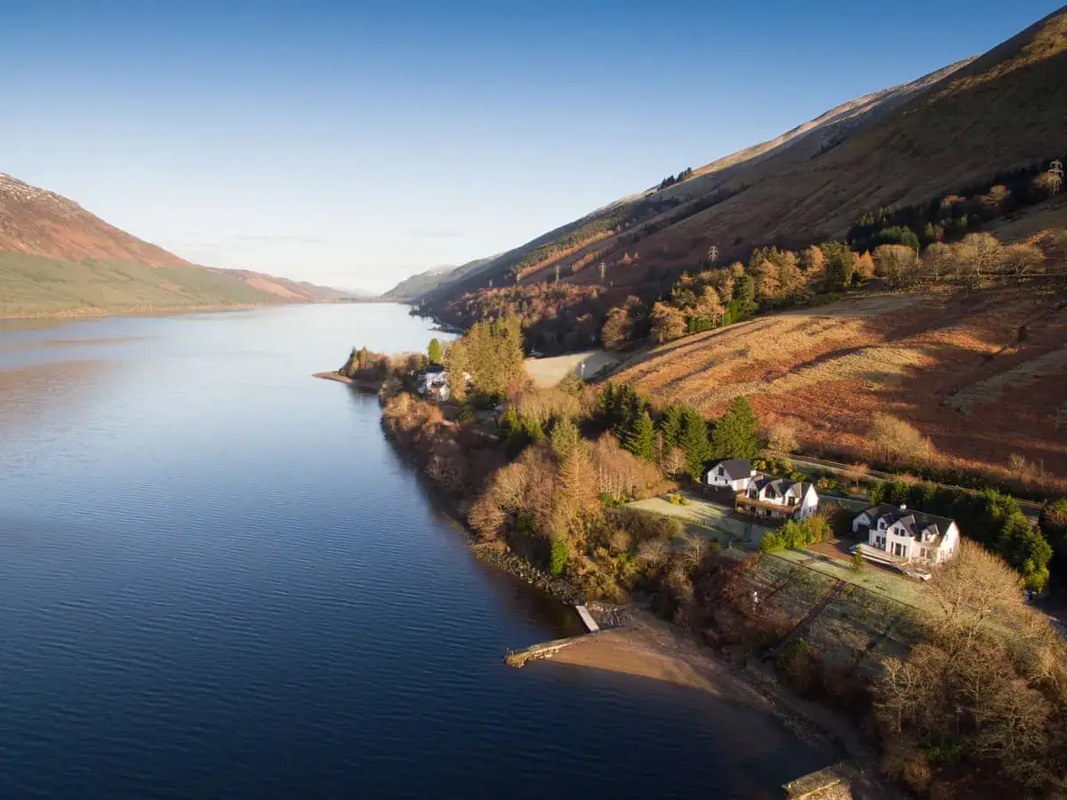 Serene gay lake with houses nestled on a wooded hillside. The sun casts long shadows across the autumnal landscape.