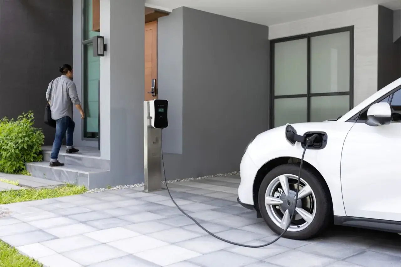 Mężczyzna wchodzi do domu, podczas gdy biały samochód elektryczny ładuje się przy domowej stacji ładowania. Sprawdź domowa stacja ładowania samochodów elektrycznych cena.