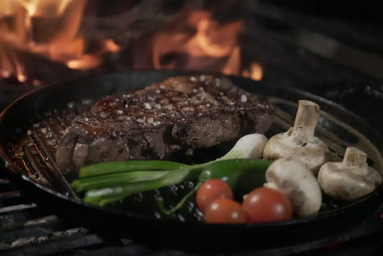 En saftig flapsteak marinad p&aring; grillen med champinjoner, tomater och gr&ouml;n chili. Flammorna dansar i bakgrunden.