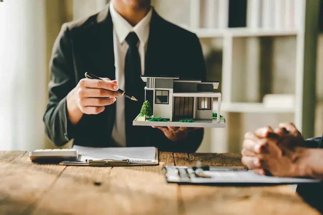 Un agente inmobiliario muestra una maqueta de casa, se&ntilde;alando los detalles mientras discute un contrato compra venta con un cliente.