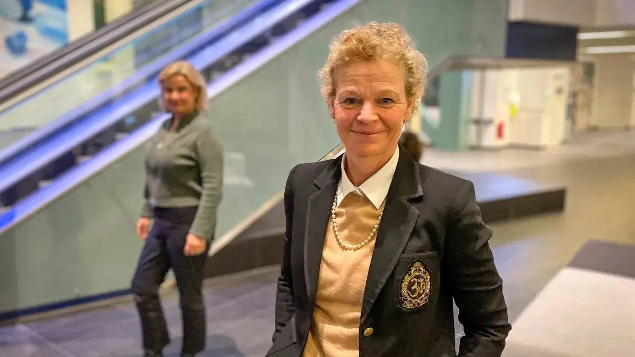 Annemarie Gardshol ler mot kameran, kl&auml;dd i kavaj och p&auml;rlhalsband. En annan kvinna syns i bakgrunden vid en rulltrappa.