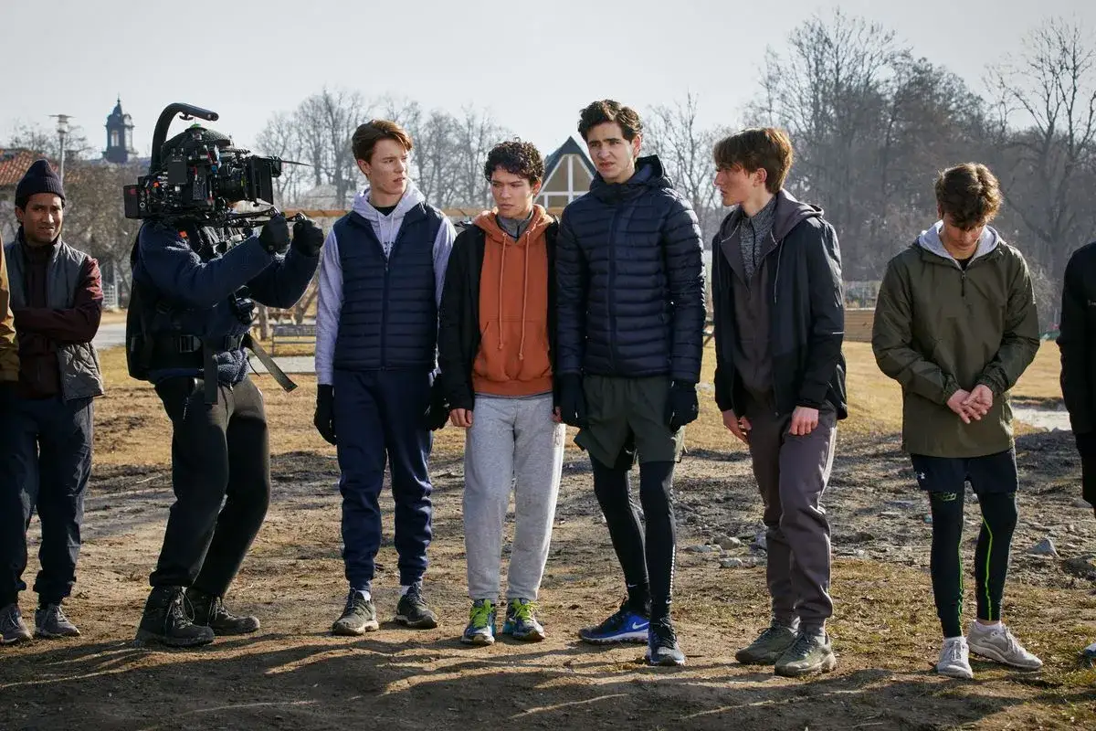 Unga sk&aring;despelare fr&aring;n Young Royals st&aring;r uppradade utomhus, en filmare med kamera syns i bakgrunden.
