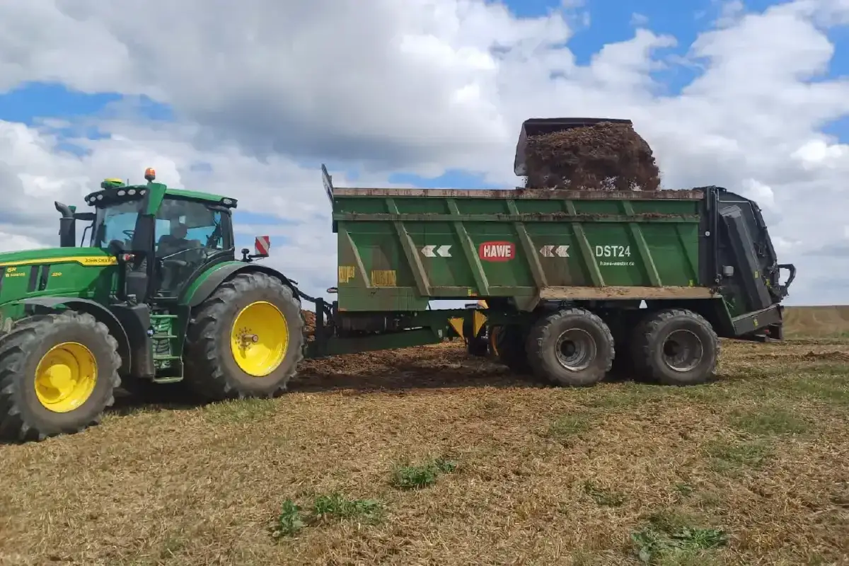 Zielony traktor John Deere z przyczepą HAWE DST24, która wysypuje obornik na pole.