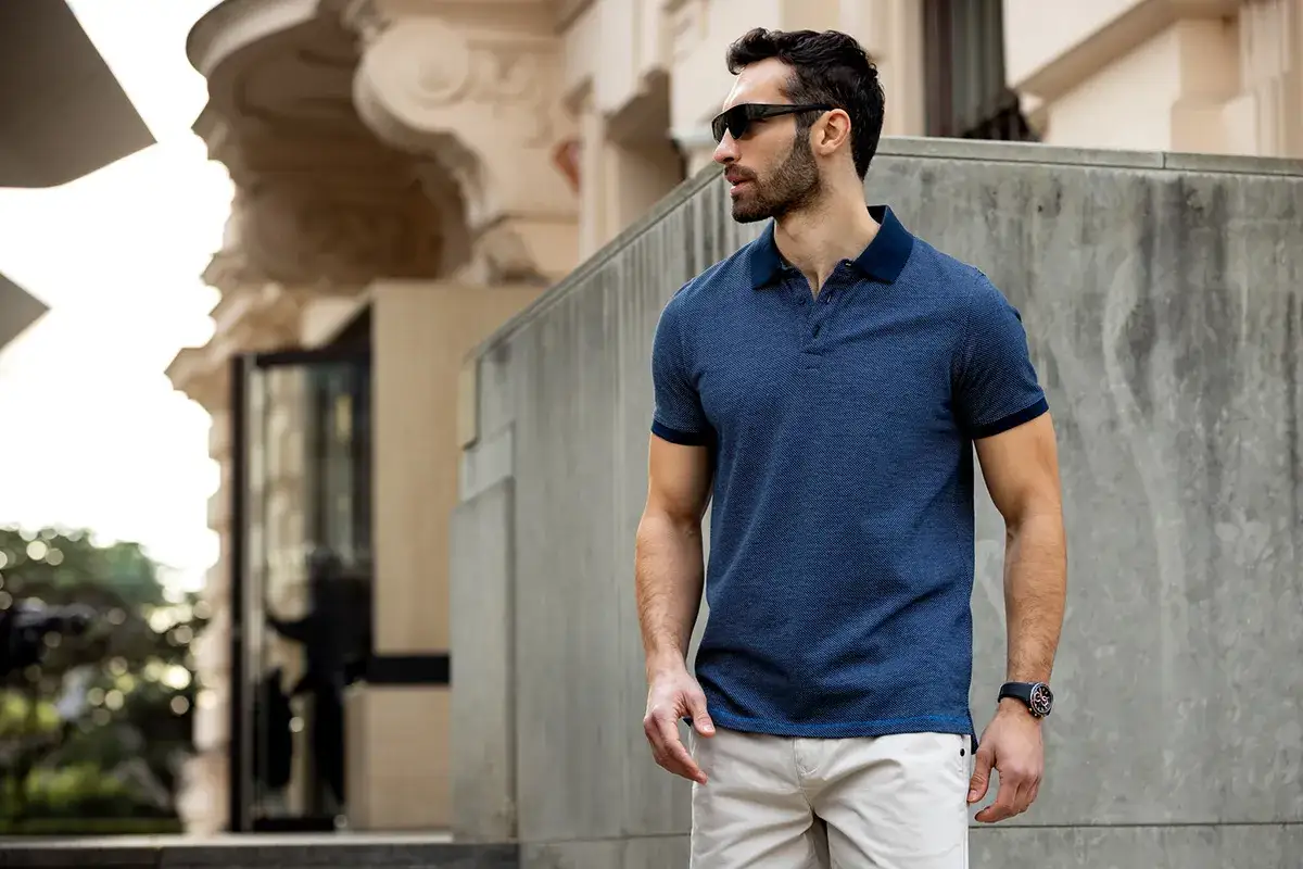 Mężczyzna w granatowej koszulce polo, która wygląda jak elegancka koszula na t-shirt, w beżowych spodniach i okularach przeciwsłonecznych.