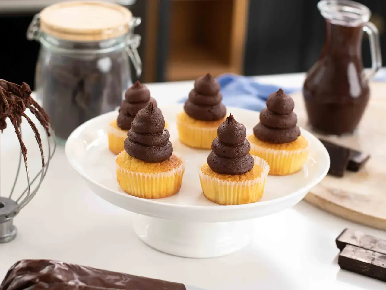 Fem cupcakes med chokladfrosting, perfekt för ett enkelt ganache recept. Ingredienser för bakning syns i bakgrunden.