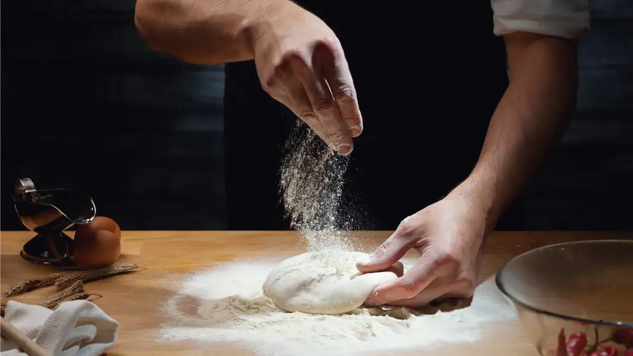 Ręce posypują mąką ciasto na pizzę, które przygotowano dzień wcześniej. Obok leżą jajka i kłosy zboża.