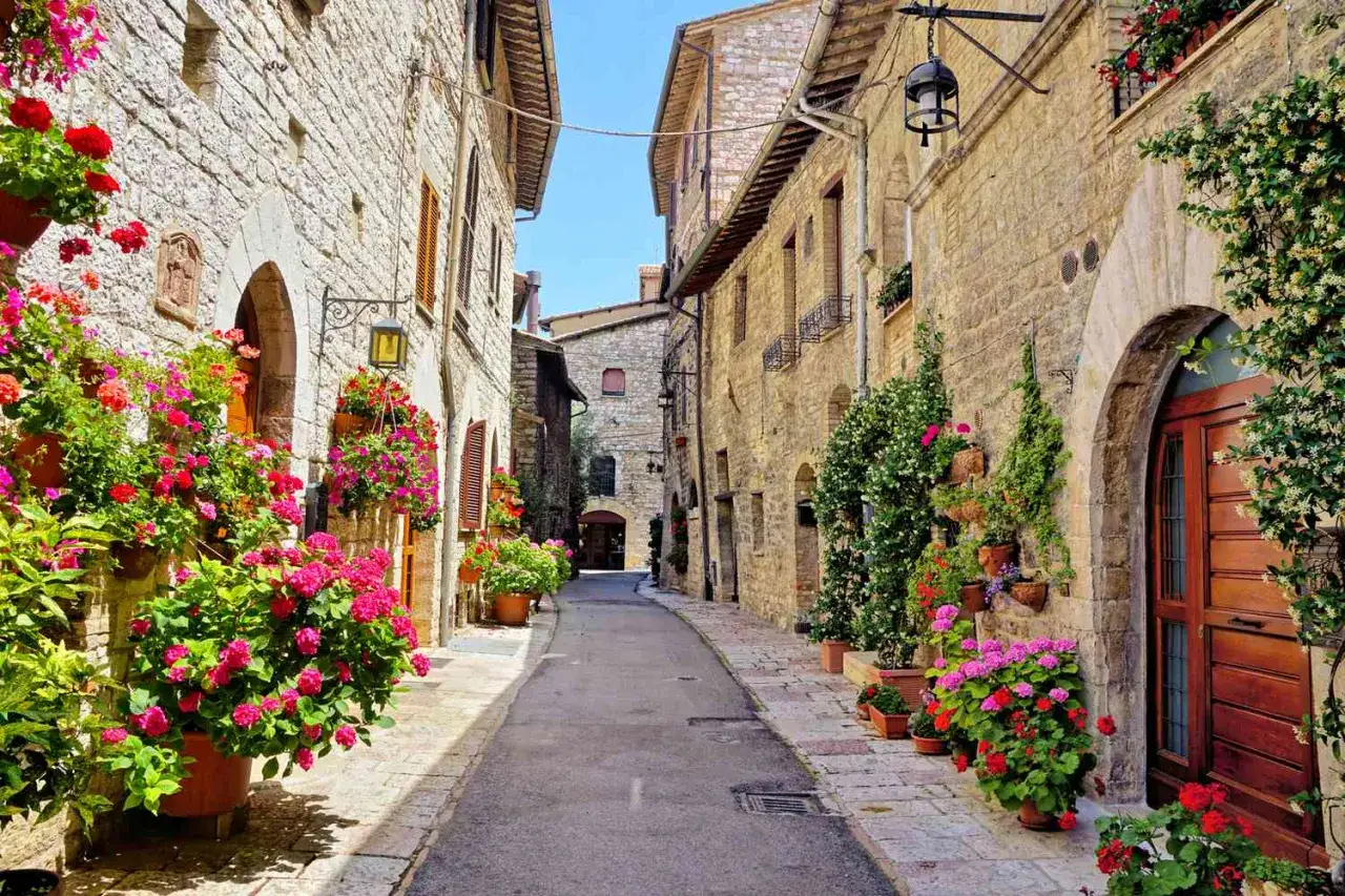 En smal gr&auml;nd i en gammal stad i Italien, kantad av stenbyggnader och &ouml;verfl&ouml;dande blommor i krukor.