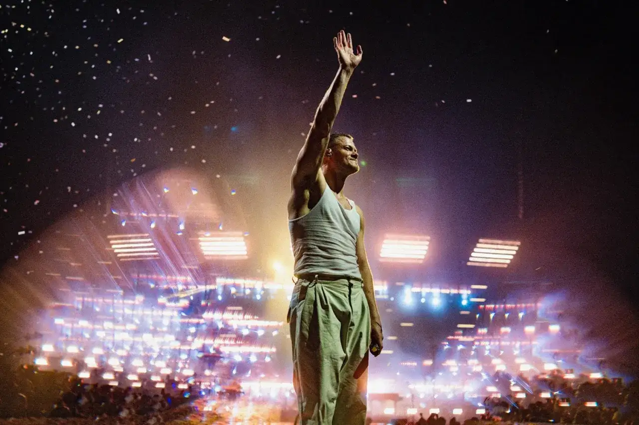 Imagine Dragons wokalista Dan Reynolds pozdrawia publiczność na koncercie. W tle migoczą światła i konfetti.