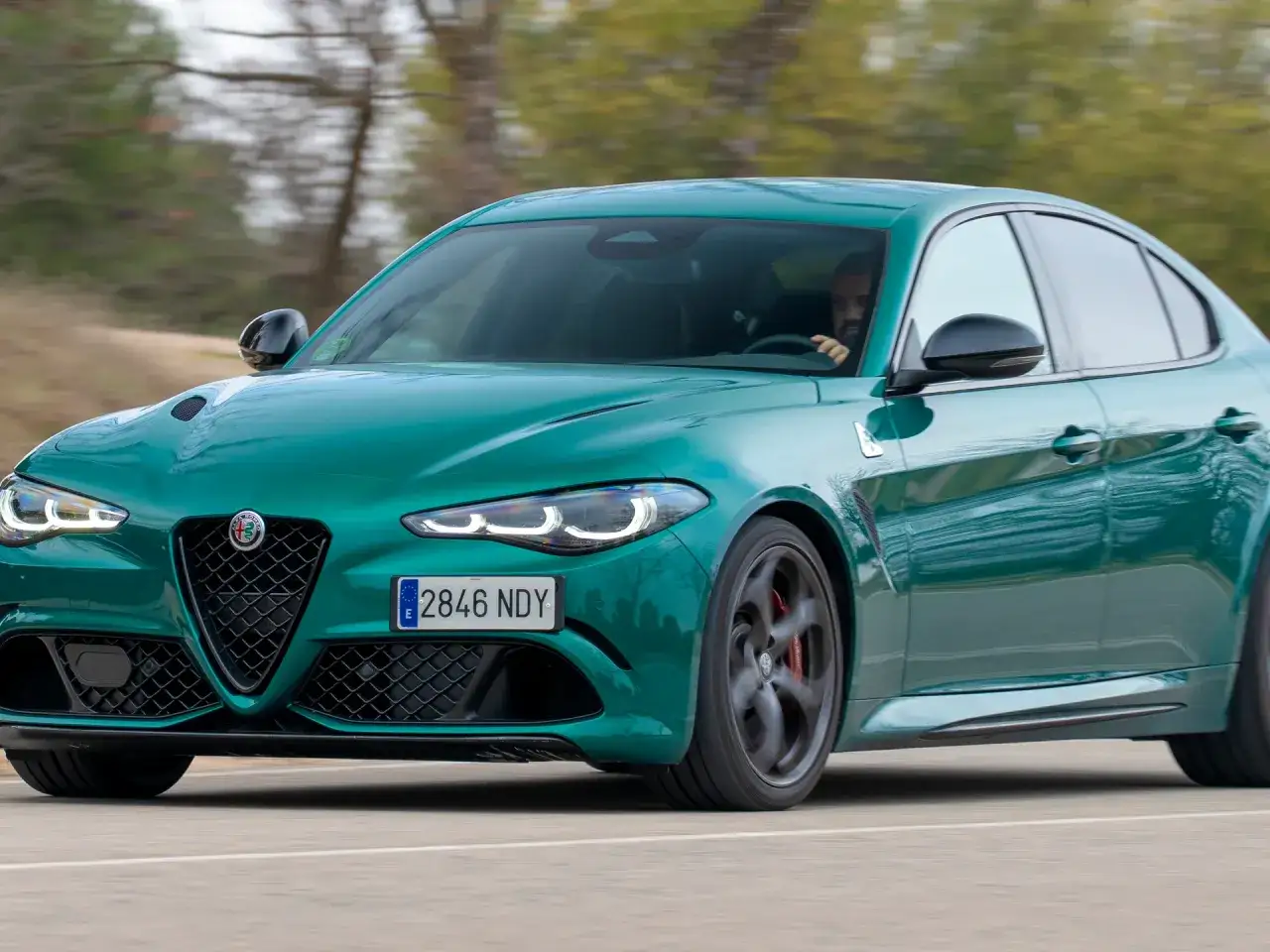 Un Alfa Romeo Giulietta Quadrifoglio Verde verde esmeralda acelera por una carretera, con el conductor al volante.