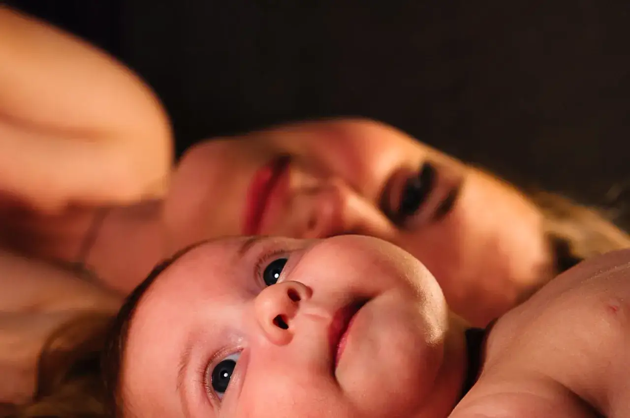 Ein Baby im REM-Schlaf, mit weit geöffneten Augen, blickt neugierig in die Kamera, während im Hintergrund die unscharfe Silhouette einer Frau zu sehen ist.
