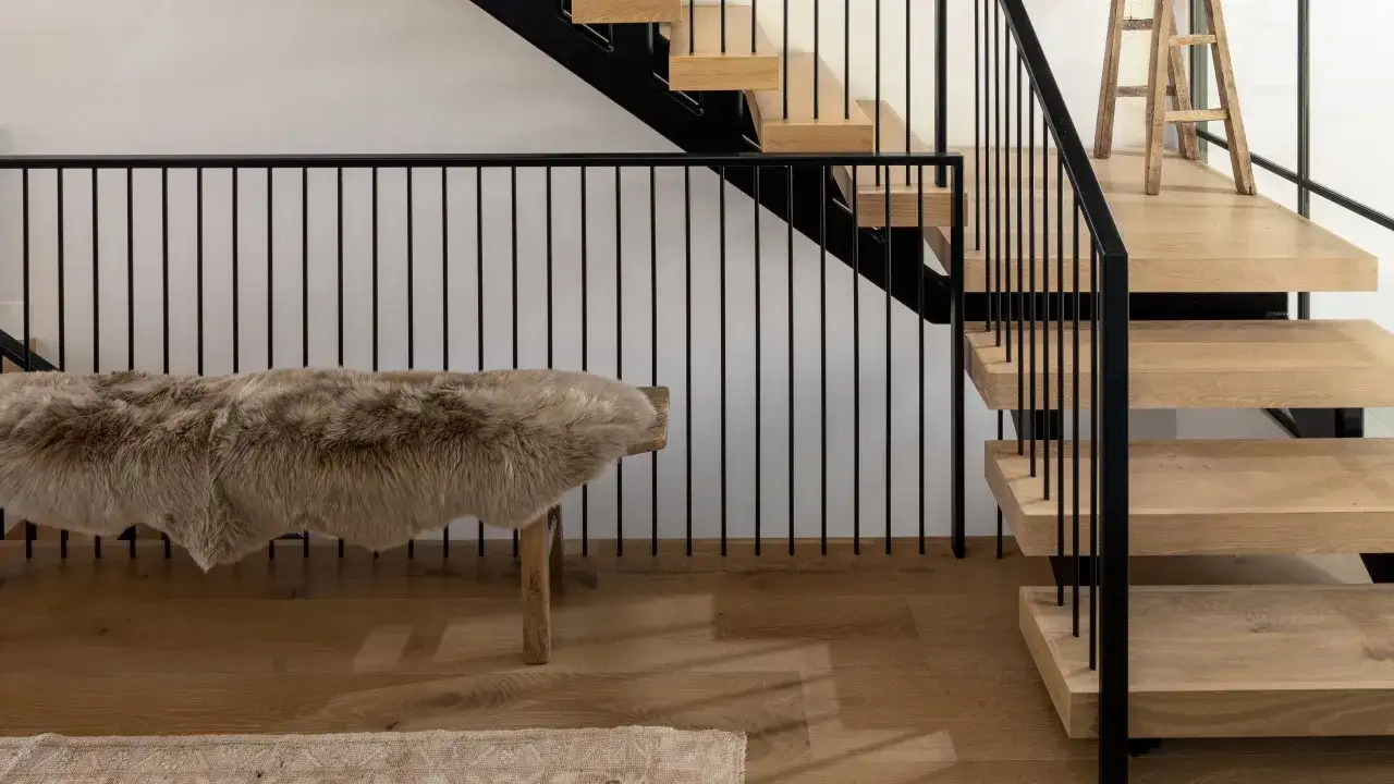 Escaleras modernas con pelda&ntilde;os de madera clara y barandilla negra. Un banco r&uacute;stico cubierto de piel a&ntilde;ade calidez.
