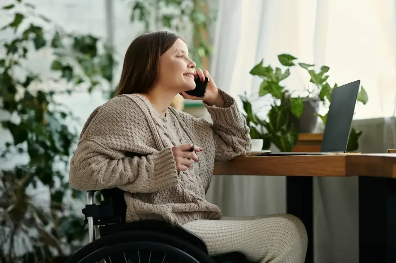 Kobieta na wózku rozmawia przez telefon, planując dofinansowanie do mieszkania. Obok laptop i rośliny.