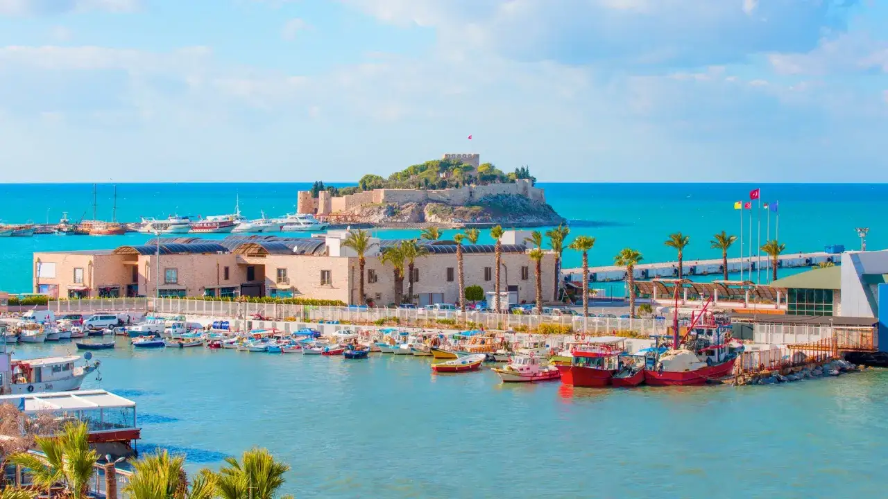 Majowa pogoda w Kusadasi zachęca do rejsu. Widok na port z zamkiem na wzgórzu i palmami.