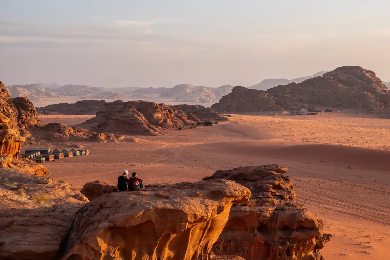 Dwie osoby siedzą na skale, podziwiając rozległą pustynię Wadi Rum o zachodzie słońca. W oddali widać ob&oacute;z i skaliste g&oacute;ry.