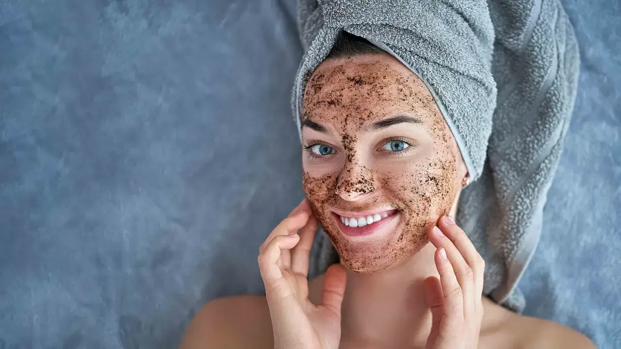 Mujer sonriente con mascarilla de café para exfoliar su rostro, envuelta en una toalla gris.
