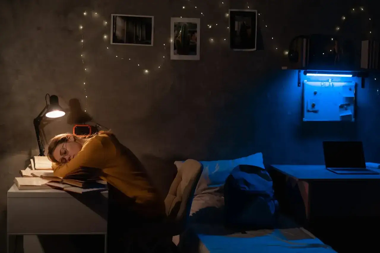 A college student sleeps at their desk, surrounded by books and fairy lights. This scene answers the question: what time do college students go to bed?